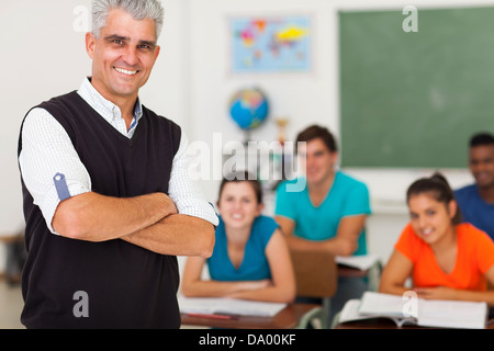 Sorridente di mezza età insegnante di scuola superiore con i bracci ripiegati in piedi di fronte alla classe Foto Stock