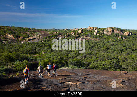 I turisti escursioni al di sopra di grande grotta Camp (a distanza) si mescola al affioramenti granitici della Matopos Hills, Zimbabwe, Africa Foto Stock