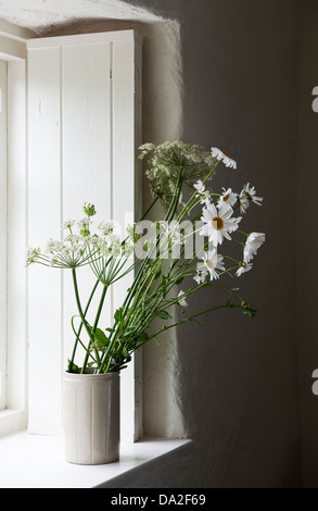 Un davanzale di cottage dipinto di bianco in estate, con persiane bianche e una semplice caraffa bianca di fiori selvatici bianchi, prezzemolo di mucca e margherite all'occhio di bue (UK) Foto Stock