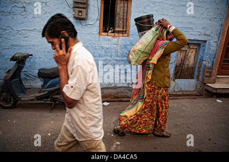 Uomo che parla al telefono cellulare e la donna borse e zaini sul suo capo in un tipico blu street nella città vecchia. Jodhpur, Rajasthan, India Foto Stock