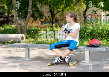 Bella ragazza in pattini mettendo in pausa per una pausa da lei pattinaggio seduta su una panchina con una grande bottiglia di plastica di acqua. Foto Stock