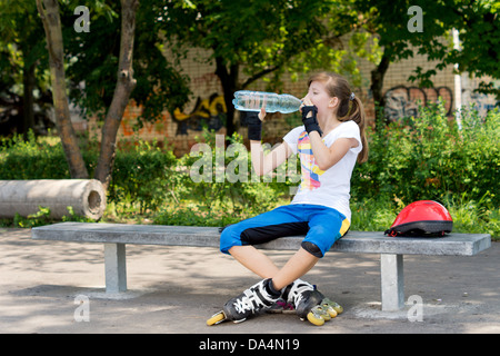 Piuttosto giovane femmina rullo teenage skater messa in pausa per un drink seduti su una panchina di bere acqua in bottiglia per placare la sua sete Foto Stock