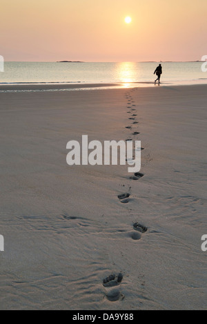 Uomo a camminare lungo la spiaggia lasciando tracce nella sabbia di sunrise. Bamburgh, Northumberland, Inghilterra Foto Stock