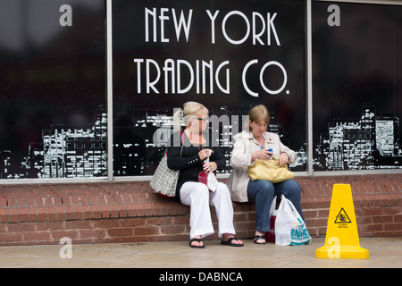 Due signore fuori shopping seduto da un 'New York Trading Co.' poster e avente una sigaretta in una cittadina inglese Foto Stock