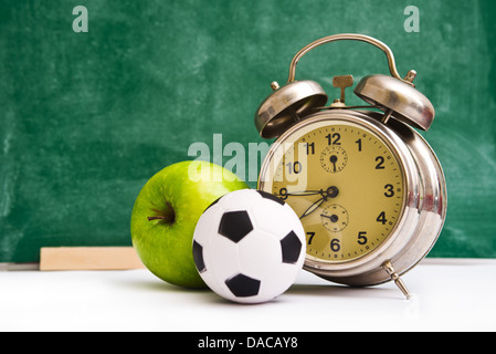 Scuola di nuovo tempo. Orologio, piccola palla e apple su insegnante del tavolo, lavagna verde in background. Si torna a scuola. Foto Stock