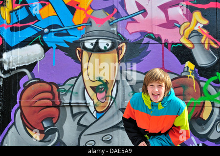Dodici-anno-vecchio ragazzo in piedi di fronte a un muro di graffiti e pocking la lingua fuori, in Germania, in Renania settentrionale-Vestfalia, Colonia Foto Stock