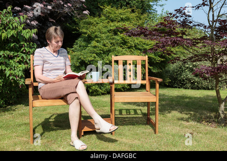 Una donna anziana in pensione si siede da sola leggendo un libro all'aperto rilassarsi su un posto a sedere in un giardino interno A casa in una giornata di sole in estate Regno Unito Foto Stock