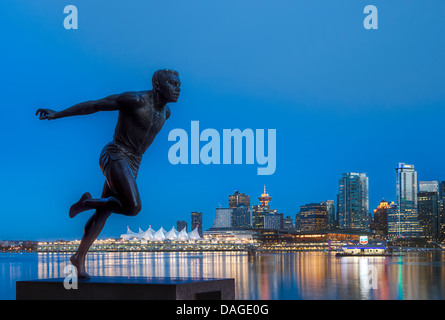 Il monumento in bronzo di runner Harry Jerome al crepuscolo, Stanley Park, Vancouver, BC, Canada. Il centro cittadino e il Canada Place attraverso Coal Harbour in background. Foto Stock