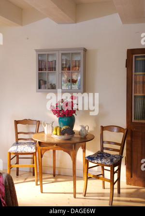 Piccolo grigio verniciato armadietto a parete al di sopra di sedie antiche e piccolo tavolo circolare con vaso di rose rosa nel paese sala da pranzo Foto Stock