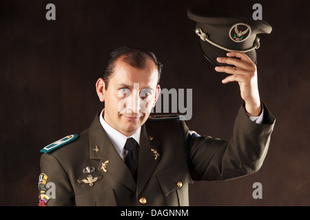 Un ufficiale militare maschile con un'uniforme verde oliva scuro si erge su uno sfondo scuro e neutro. Sta tenendo il berretto di servizio nella mano destra, sollevato con un gesto di saluto vicino alla testa. Il Foto Stock