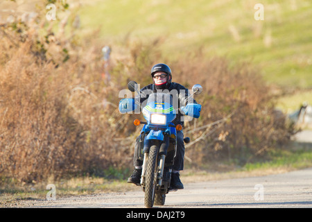 Vecchio uomo che indossa un casco e occhiali protettivi in sella a una motocicletta (cross bike) su una strada attraverso un parco Foto Stock