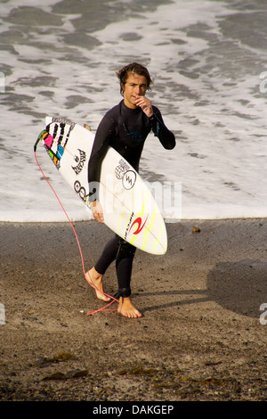 Indossare muta e trasportare le tavole da surf, ragazzo adolescente membro dell'alta scuola surf team emerge dall'Oceano Pacifico. Foto Stock