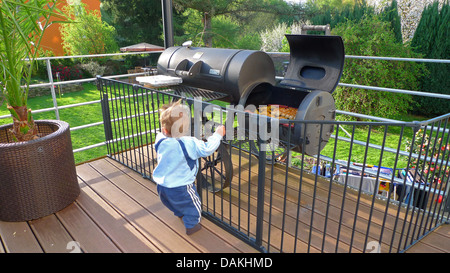 Little Boy curiosamente in piedi in un recinto su una veranda davanti a un grill pieno di carne e salumi Foto Stock