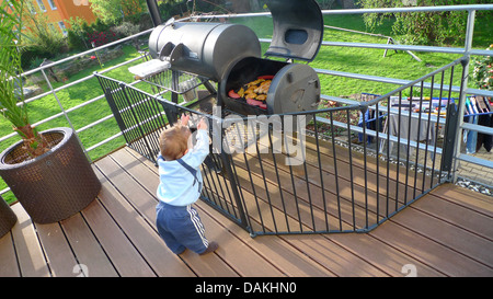 Little Boy curiosamente in piedi in un recinto su una veranda davanti a un grill pieno di carne e salumi Foto Stock