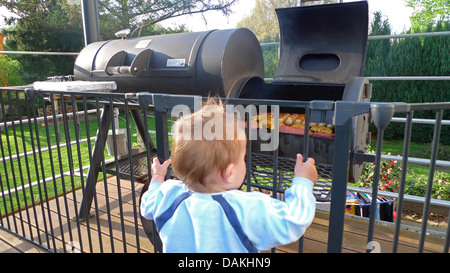 Little Boy curiosamente in piedi in un recinto su una veranda davanti a un grill pieno di carne e salumi Foto Stock