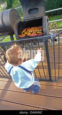 Little Boy curiosamente in piedi in un recinto su una veranda davanti a un grill pieno di carne e salumi Foto Stock