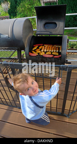 Little Boy curiosamente in piedi in un recinto su una veranda davanti a un grill pieno di carne e salumi Foto Stock