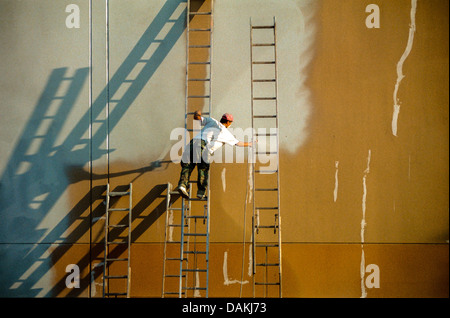 In equilibrio su una scala, un operaio ispanico vernici spray di un muro di fabbrica a Irvine, CA. Nota lunghe ombre dal sole del pomeriggio. Foto Stock