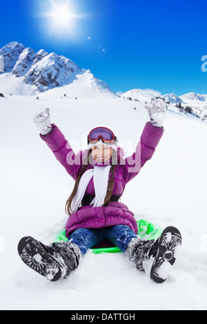 Felice ragazza scorrevole sulla slitta con le mani sollevate, indossa una maschera da sci, con le montagne sullo sfondo Foto Stock