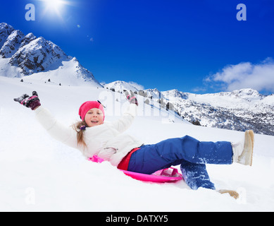 Felice ragazza scorrevole sulla slitta con le mani sollevate, indossa una maschera da sci, in montagna Foto Stock