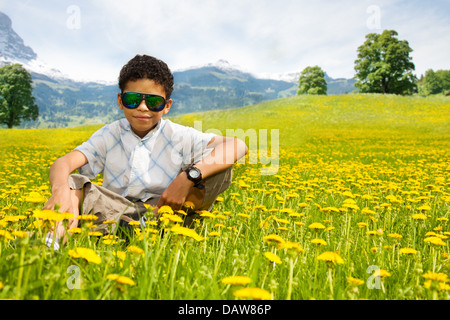 Happy little black boy in occhiali da sole seduti nel campo di tarassaco con le montagne sullo sfondo Foto Stock