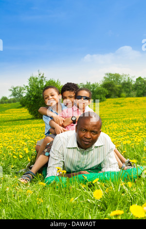 Nero uomo africano con ragazzi seduto sul suo retro recante nella primavera del parco con campo di tarassaco giallo Foto Stock