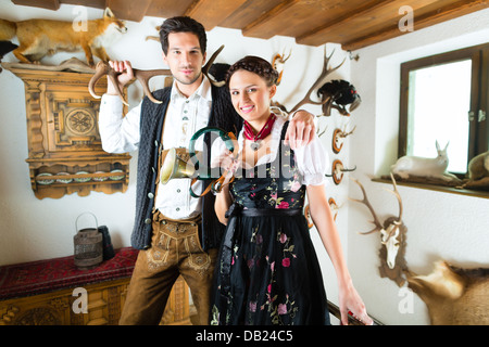 Giovane cacciatore con sua moglie e una tromba davanti a una parete con diversi corna, palchi e trofei in una baita alpina Foto Stock