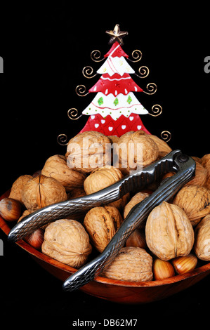 Ciotola di legno pieno di noci e nocciole con schiaccianoci e un grazioso stagno albero di Natale per la parte posteriore su sfondo nero. Foto Stock