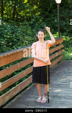 Foto verticale della ragazza che regge trote piccole, mentre la pesca off ponte di legno con alberi e lampada posta in background Foto Stock