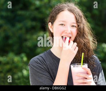 Foto della ragazza giovane mangiare fragole intere tenendo frullato in altra parte sfocata fuori luminose verdi alberi in backgro Foto Stock