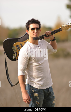 Un musicista rock indossando occhiali da sole portano brown chitarra elettrica sulla spalla sinistra in posa all'aperto in natura caduta meteo Foto Stock