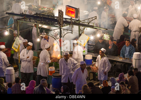 Affacciato su un alfresco stallo alimentare che mostra i cuochi che circonda una cottura fumoso fuoco, Djemaa el-Fna di notte a Marrakech, Marocco Foto Stock