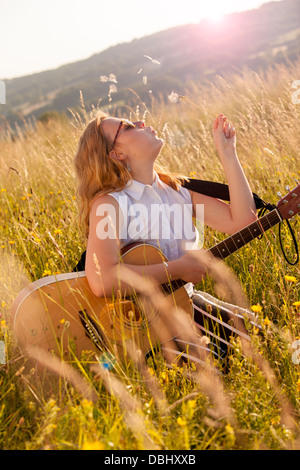 Ragazza adolescente indossando occhiali da sole scuri seduto in un campo erboso con una chitarra nel suo giro soffiando a un seme dandilion testa. All'esterno. L'estate. Foto Stock