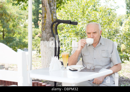 Anziani disabili uomo gustando una tazza di caffè e dessert a un open-air cafe con sue stampelle appoggiato sul tronco di albero accanto a lui. Foto Stock