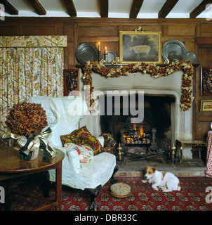 Terrier cane sdraiato sul tappeto davanti al fuoco nel paese soggiorno con secchi fiori e ala bianca sedia Foto Stock