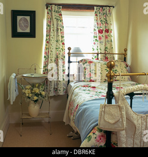 Rose-tende a motivi geometrici sulla finestra sopra il letto in ottone con abbinamento di rose-stampa di biancheria da letto nella camera da letto cottage con lavabo vintage Foto Stock
