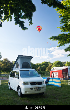 Un colorate mongolfiere volare al di sopra di un campeggio con e una VW camper van piantato su di una tranquilla serata d'estate. Foto Stock