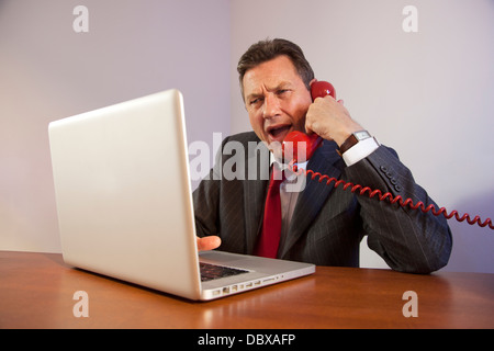 Angry Man indossa una tuta, gridando verso il basso un telefono rosso seduti davanti a un computer portatile su una scrivania. Foto Stock