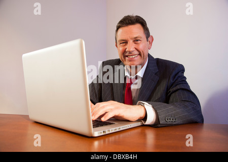 Uomo d'affari a una scrivania utilizzando un computer portatile, guardando la telecamera sorridendo. Foto Stock