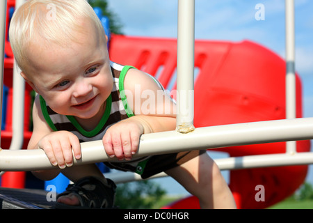 Un simpatico, happy baby boy è sorridente come egli si accovaccia giù e tenta di attraversare un ponte ad un parco giochi in un giorno di estate Foto Stock