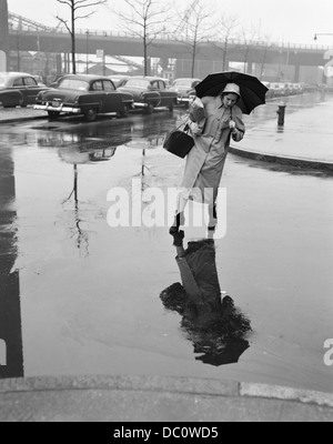 Anni Cinquanta la donna in cappotto di pioggia HAT STIVALI HOLDING OMBRELLO STEPPING BORSETTA PER EVITARE pozzanghere durante l attraversamento strada di città in fallo METEO Foto Stock