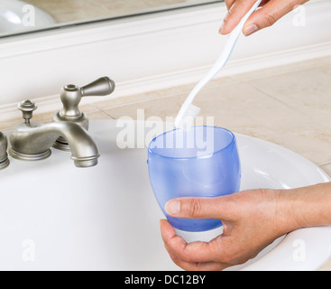 Primo piano delle mani femminili mettendo spazzolino da denti in blu cup con bagno bianco lavello e rubinetto in background Foto Stock