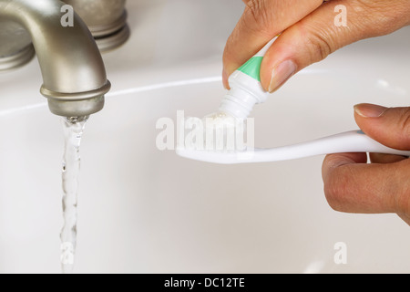 Ingrandimento di foto orizzontale di mani femminili di mettere pasta dentifricia su uno spazzolino da denti con bagno bianco lavello e rubinetto in esecuzione in bac Foto Stock