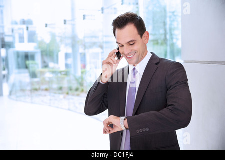 Imprenditore sorridente parlando al cellulare e guardando un orologio da polso Foto Stock