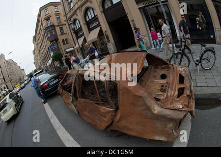 I passanti da guardare a un'auto bruciata, che era stata parcheggiata su una striscia di parcheggio come opera d'arte, sulla Maximilianstrasse a Monaco di Baviera, Germania, 07 agosto 2013. Sotto l'auto rottamata un telaio metallico con piccole ruote è montata, e dall'interno frammenti di una conversazione può essere sentito. Foto: PETER KNEFFEL Foto Stock