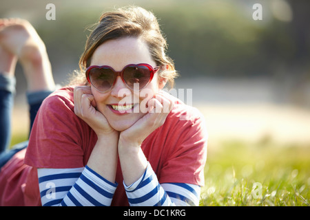 Ritratto di giovane donna che indossa forma di cuore occhiali da sole sdraiati sull'erba Foto Stock