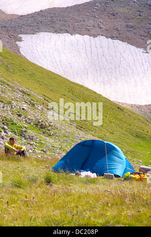 Tenda da campeggio in montagna con uomo escursionista seduto su erba e Glacier Hillside Su sfondo Foto Stock