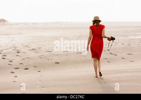 Donna matura indossando abito rosso camminando sulla spiaggia Foto Stock