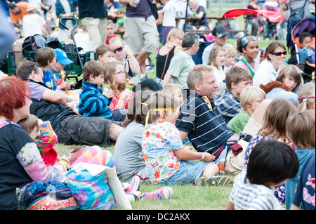 Una folla di bambini felici, adulti e famiglie seduto sull'erba godendo di un festival estivo di guardare un animatore di bambini Foto Stock