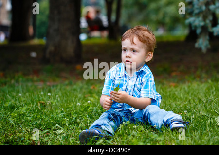 Little Boy in jeans e un plaid shirt seduto sull'erba nel parco Foto Stock